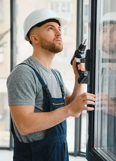 A construction worker in a hard hat and overalls using a drill on a window frame.