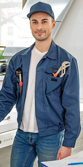 A man in a blue work uniform and cap, smiling, with tools in his jacket pocket, standing indoors.