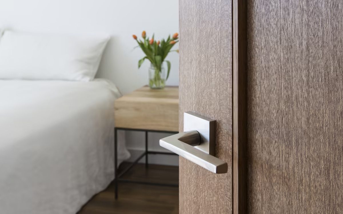 Close-up of a modern metal door handle on a wooden door, partially opened to reveal a bedroom with a white bed and side table with orange flowers.