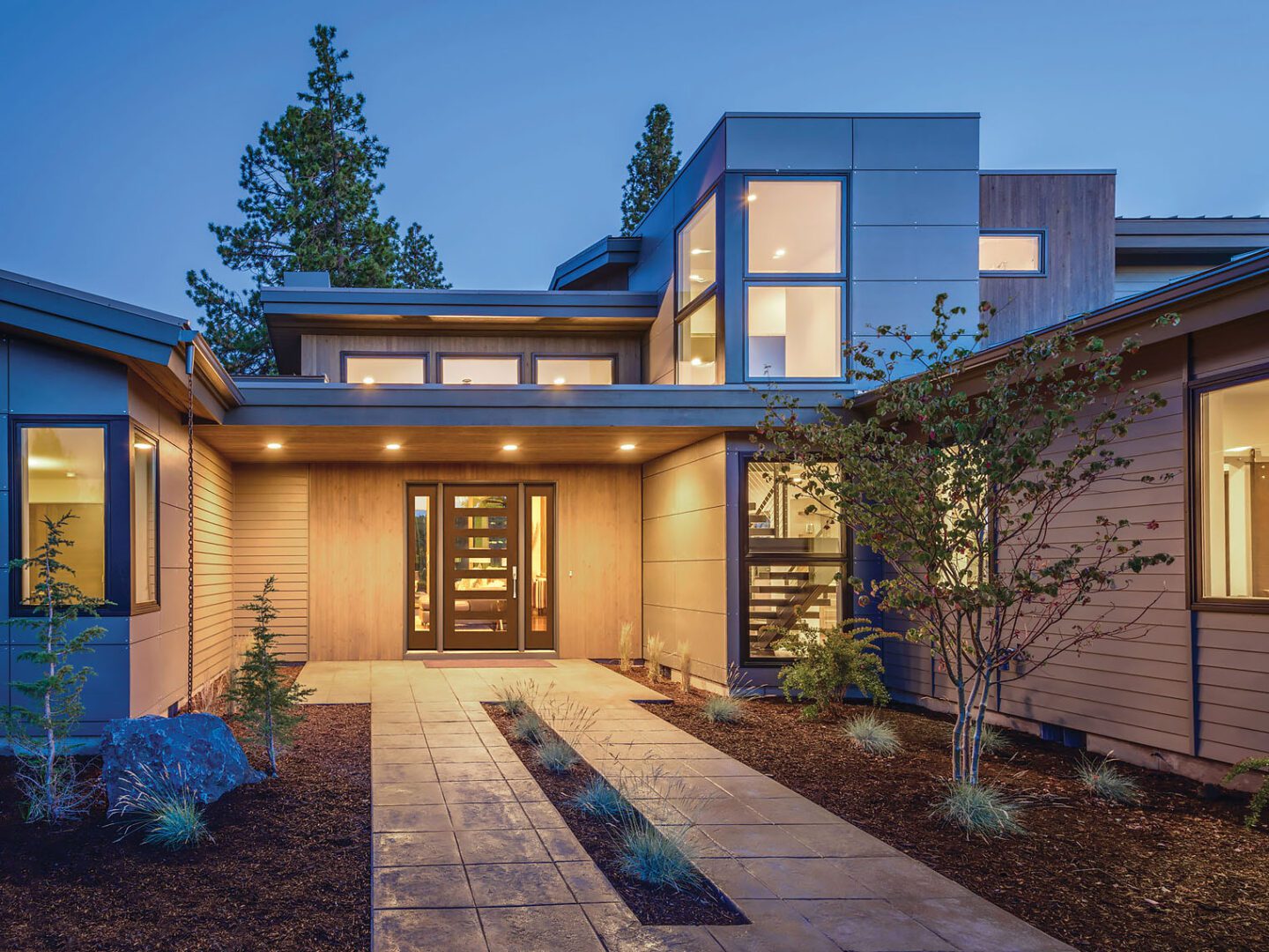 Modern house with illuminated windows at dusk, featuring a pathway leading to a wood-paneled entrance.