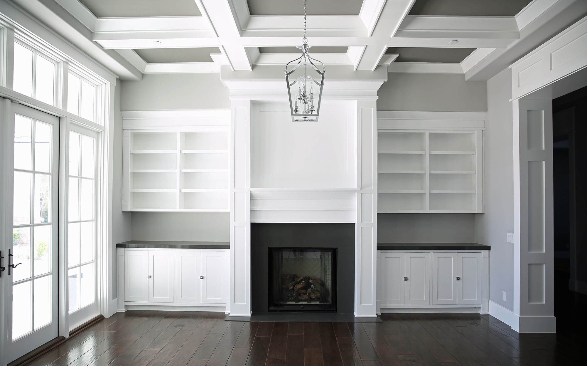 A spacious living room with white built-in shelves, a fireplace, coffered ceiling, and dark hardwood floors, with sunlight streaming through a french door.