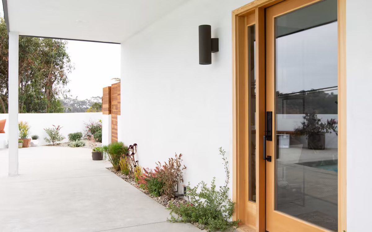 Modern house front with wooden door, white walls, a covered patio area, and potted plants.