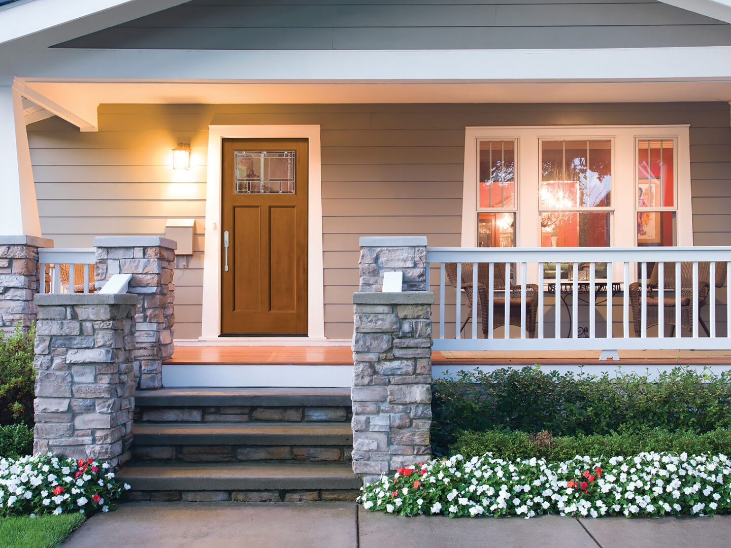 A cozy suburban home at dusk, featuring a wooden front door, stone pillars, a porch with railings, and blooming flowers along the pathway.